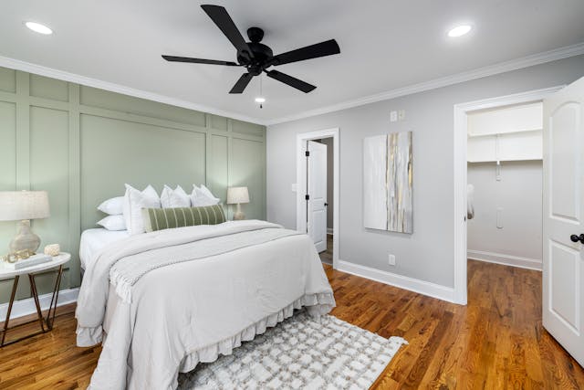 bedroom with a white bed green accent wall nightstands with lamps a ceiling fan and wooden
 flooring there is an open door leading to a walk in closet and a door to another room
